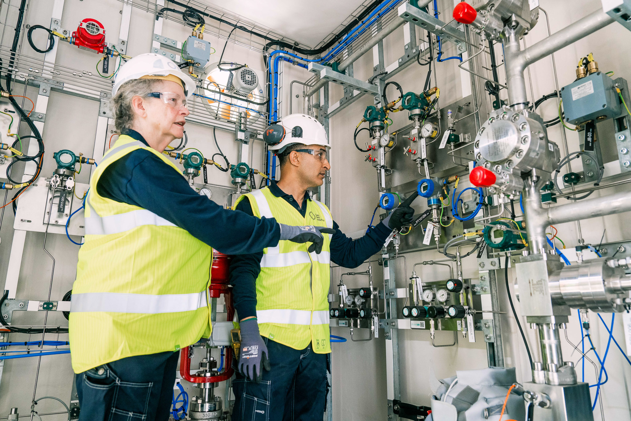 Two technicians wearing hard hats, safety glasses, and high-visibility vests inspect and point to complex industrial piping and control panels inside a facility