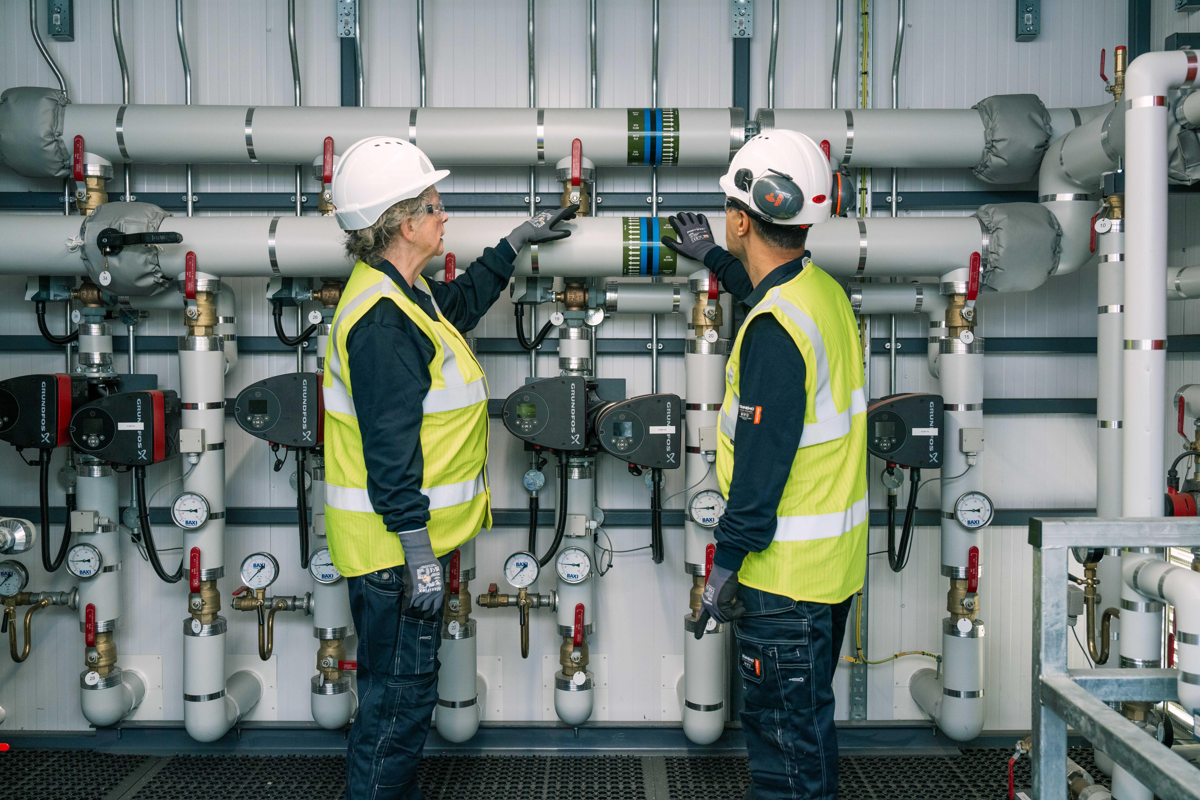 Two technicians wearing white hard hats, protective gloves, safety glasses, and high-visibility yellow vests inspect a complex industrial piping and pump system