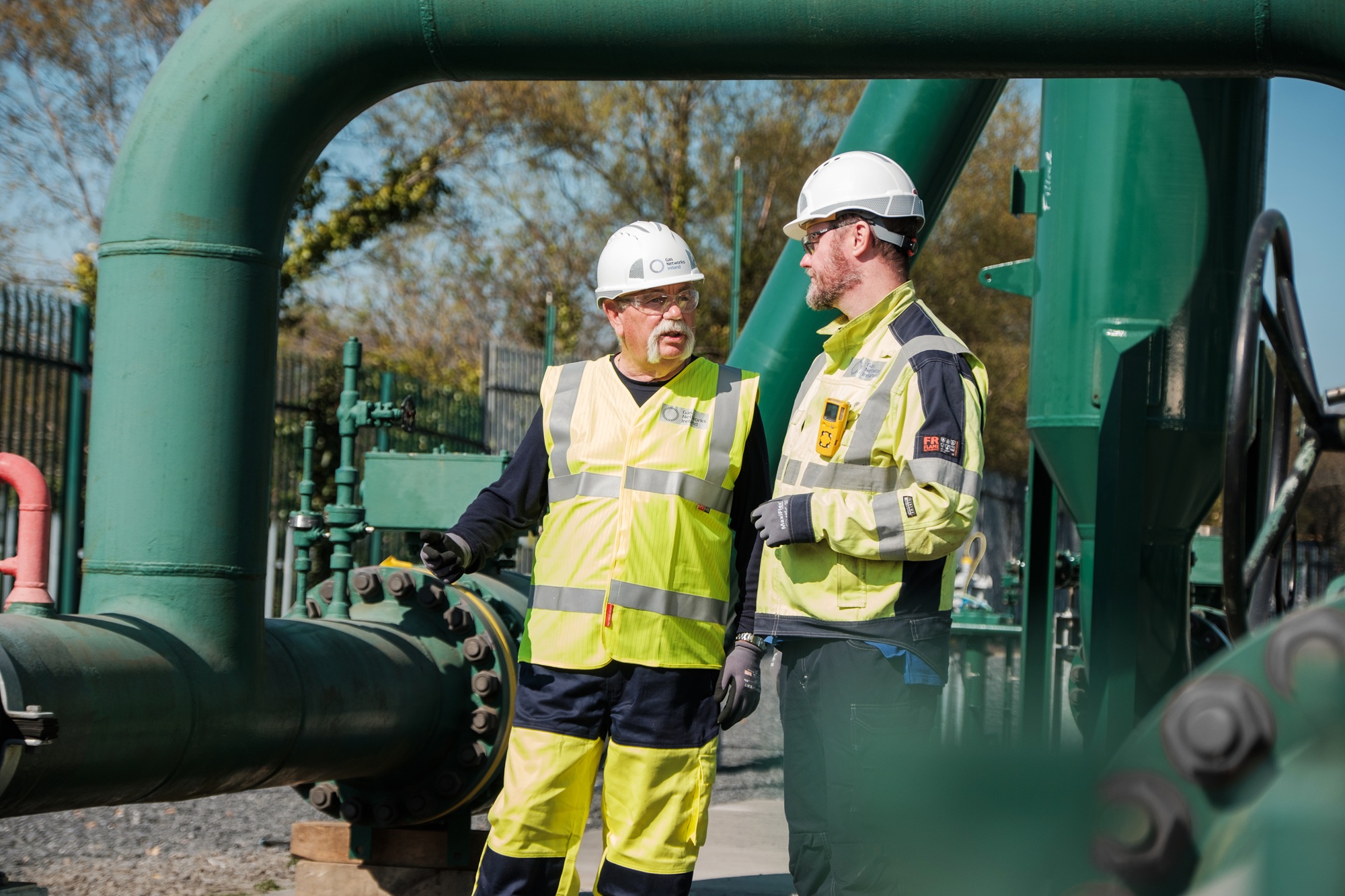 Two technicians wearing hard hats and high-visibility safety gear converse near large green industrial gas pipes outdoors.