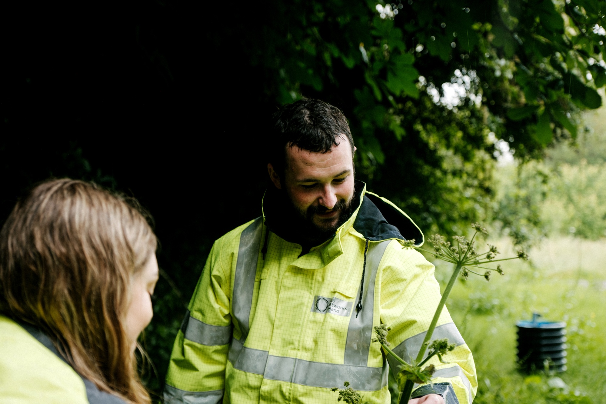 A Gas Networks Ireland worker in a high-visibility jacket examines local flora outdoors alongside a colleague during a field project.